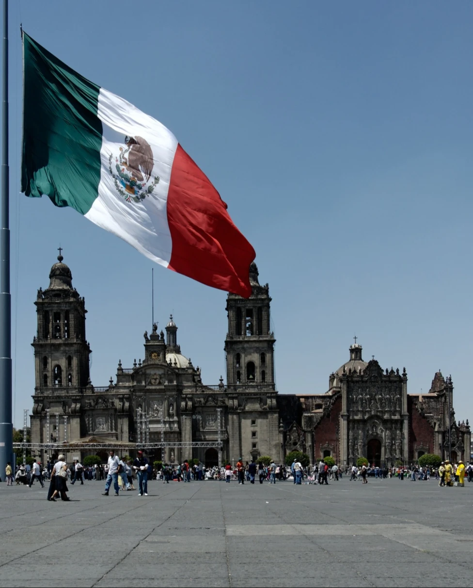 A view of the flag in Mexico in front of people walking near a grand stone building. 