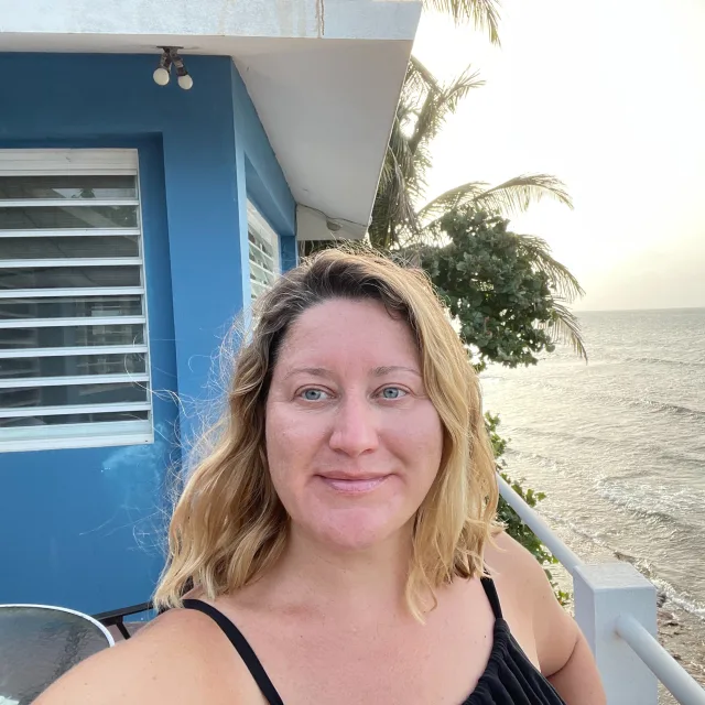 Travel Advisor Karyn Pavich in a black dress in front of a blue house and a beach.