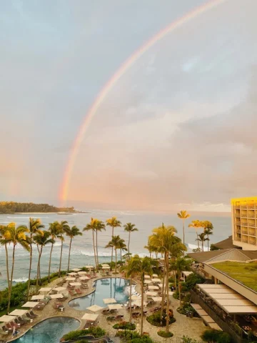 A picture of a beautiful rainbow shining over a bay with a hotel patio, pool, lounge chairs and palm trees in the forefront of the view. 