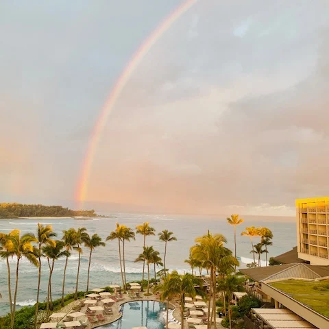 A picture of a beautiful rainbow shining over a bay with a hotel patio, pool, lounge chairs and palm trees in the forefront of the view. 