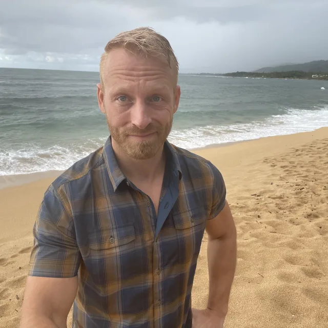 Travel Advisor Andrew Bergeron stands on a beach wearing a blue and brown plaid shirt