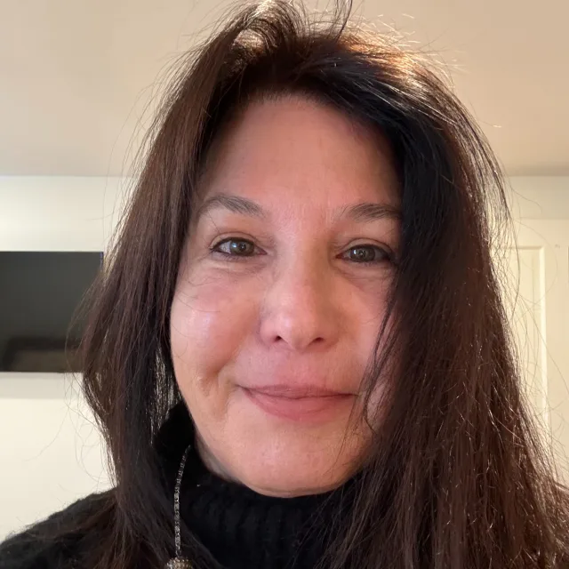 Debra smiling and wearing a black turtleneck while taking a selfie in front a white wall 