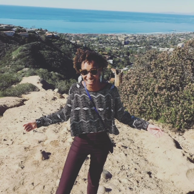 a woman in purple pants and sunglasses stands on a hill overlooking a desert valley
