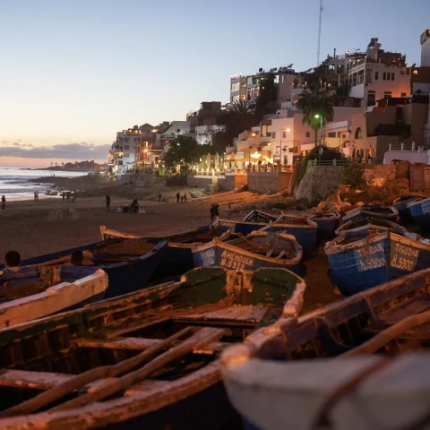 A view of boats on the beach surrounded by sand, buildings in the distance and the sea in the evening. There are people walking along the shoreline and a faint sunset in the distance. 