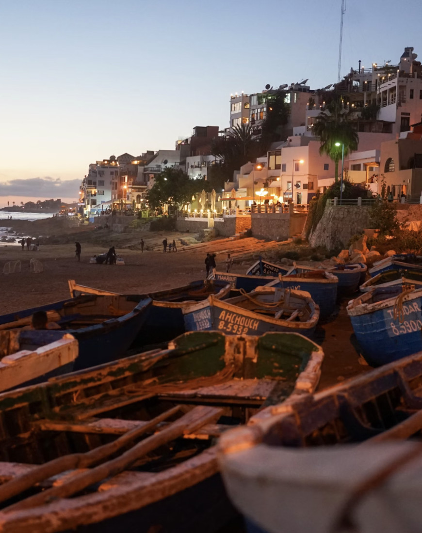 A view of boats on the beach surrounded by sand, buildings in the distance and the sea in the evening. There are people walking along the shoreline and a faint sunset in the distance. 