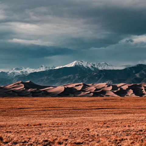 The Great Sand Dunes of Colorado from across a dim-lit field with snow-capped mountains in the far distance and heavy cloud cover (photo by Colin Lloyd)
