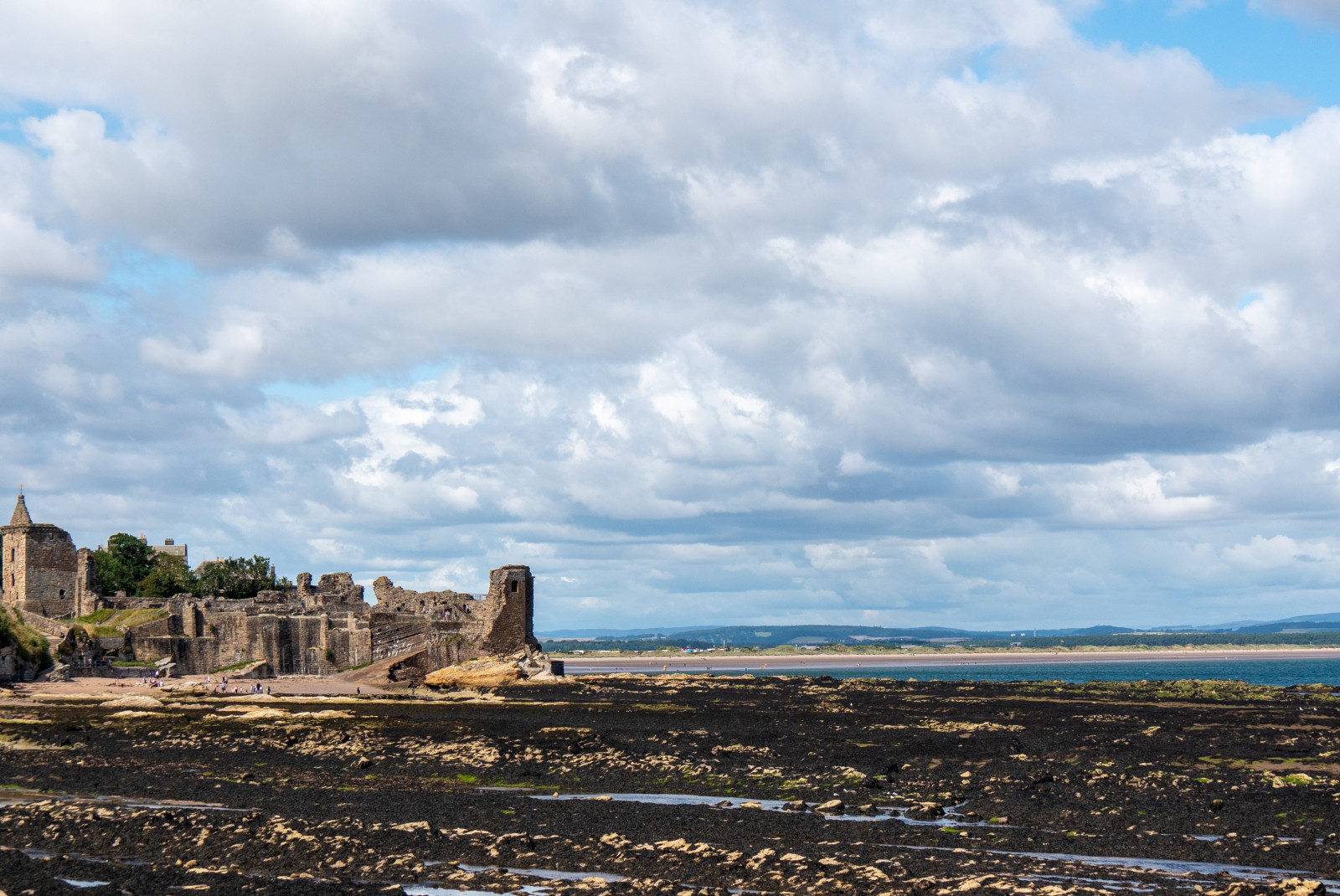 Tan St. Andrews castle ruins on a black sand beach with blue water and green hills in the distance
