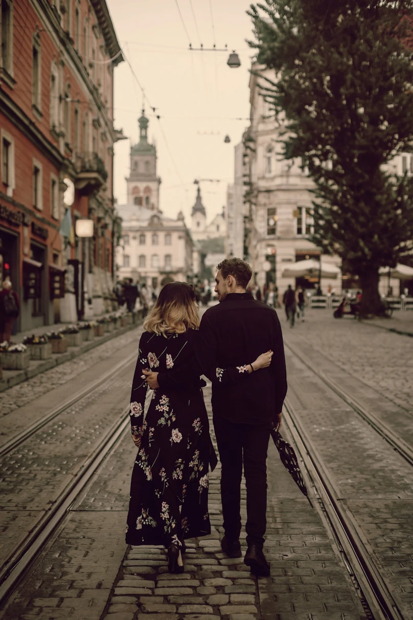 A couple strolls down a historic city street, flanked by European architecture and tram lines, immersed in the charm of their surroundings.