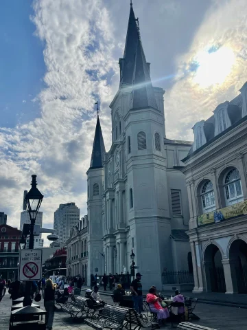 A church with tall steeples and benches in front of it with the sun peeking through clouds behind it.
