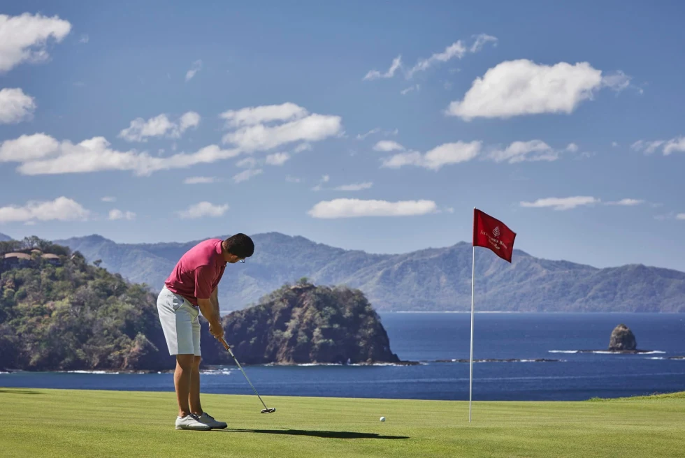 man wearing coral shirt and white shorts playing golf with mountains in the distance