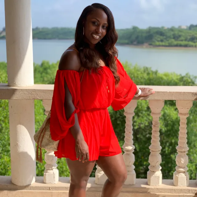 Yasmir Williams in a red dress posing on a balcony overlooking green trees and a body of water