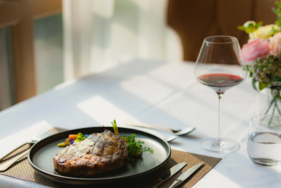 Steak on black plate next to glass of red wine and white tablecloth