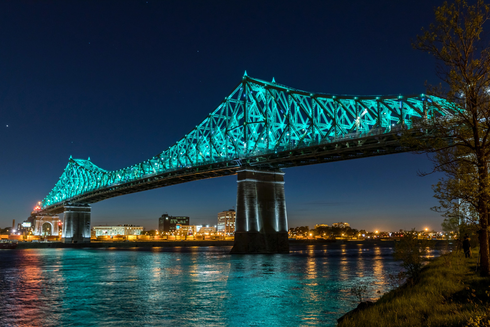 Jacques Cartier bridge in Montreal Canada lit up blue at night reflecting in a dark river and blue sky with yellow city lights in the distance