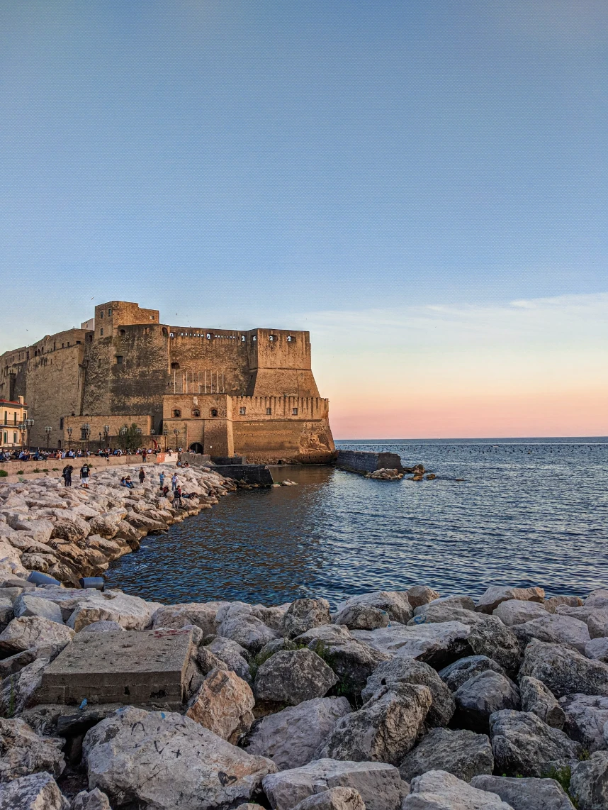 A rocky shore looking out to a rippling blue sea with a large stone fort in the background against a light pink sunset. 
