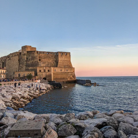 A rocky shore looking out to a rippling blue sea with a large stone fort in the background against a light pink sunset. 