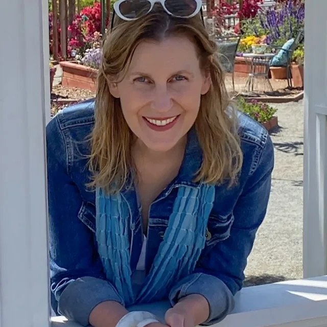 Fora travel agent Cara Todd wearing jean jacket, blue scarf and white sunglasses smiles with flowers in the background