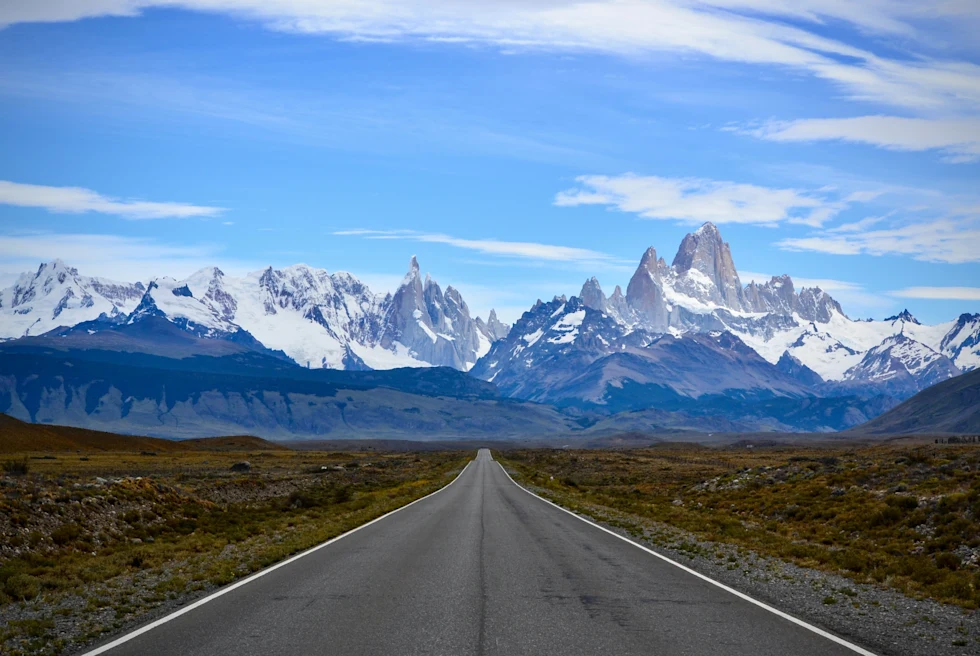 A grey concrete road leading to snow-covered mountain.