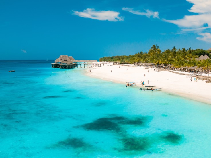 Crystal blue waters with darker patches a white sand beach green palm trees with people in the distance and a tan hut over the water 