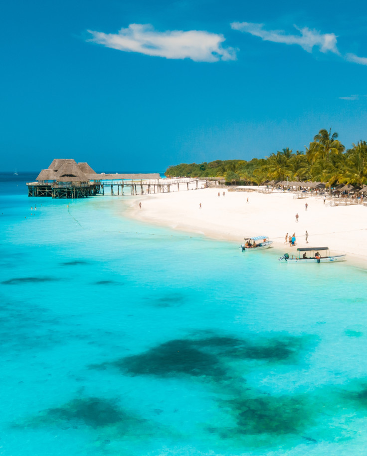 Crystal blue waters with darker patches a white sand beach green palm trees with people in the distance and a tan hut over the water 