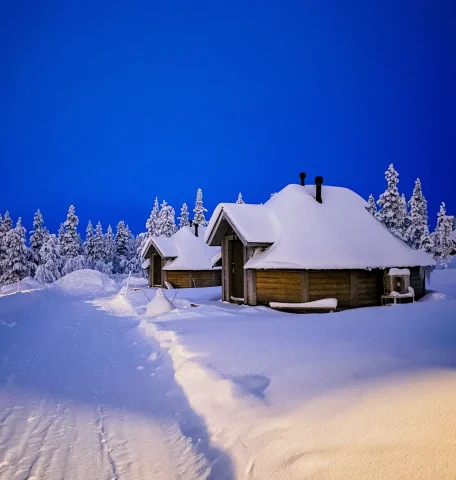 This image depicts a snow covered wooden lodge surrounded by thick and snowy terrain with pine trees in the background and a vibrant blue sky. 