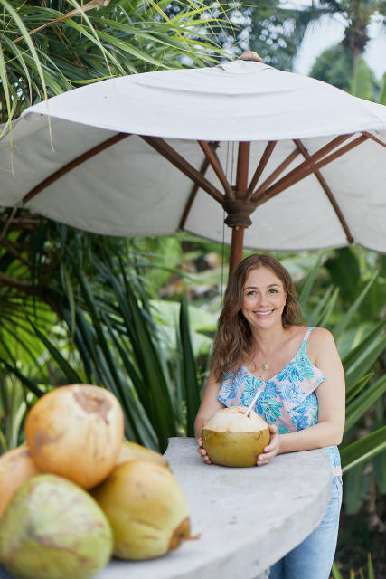 Fora travel agent Sara Beecher wears a blue shirt and holds a coconut while standing under white umbrella