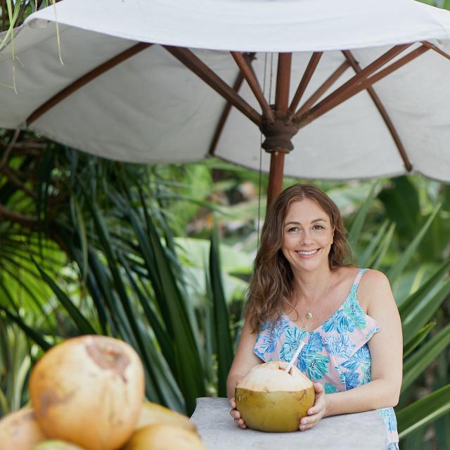 Fora travel agent Sara Beecher wears a blue shirt and holds a coconut while standing under white umbrella