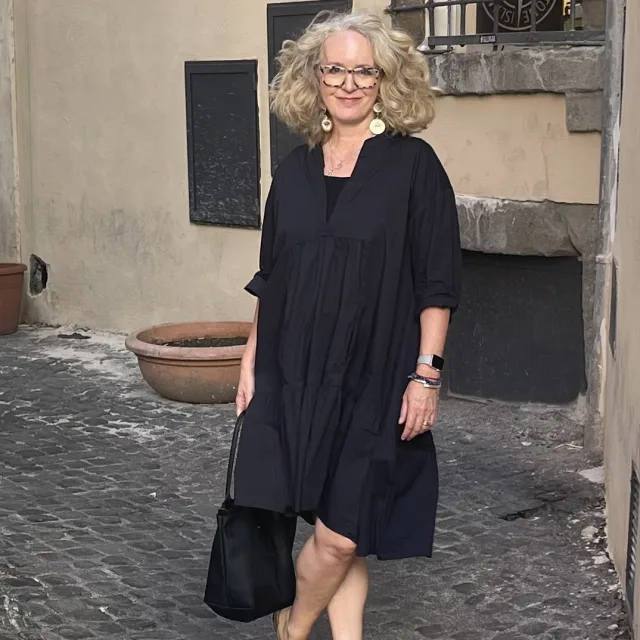 woman in glasses and a long black dress smiling on a cobblestones street