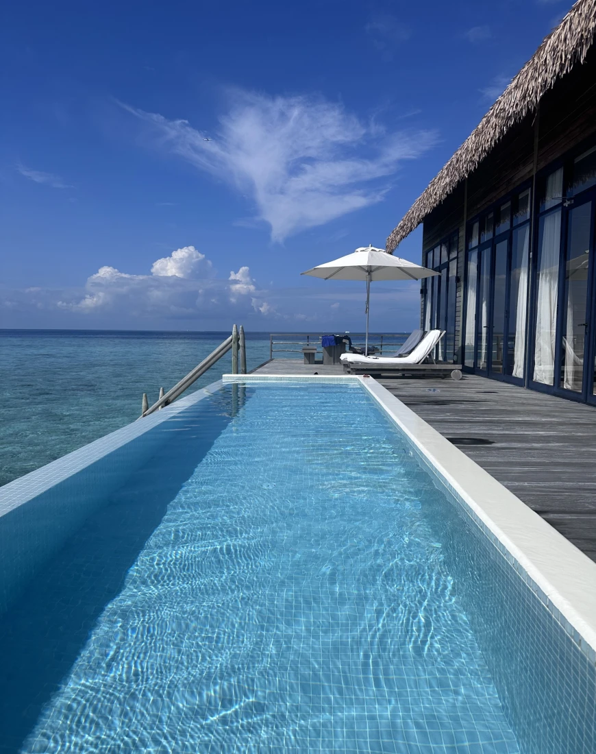 A long swimming pool next to a deck with an umbrella and lounge chair in the distance next to the sea. 