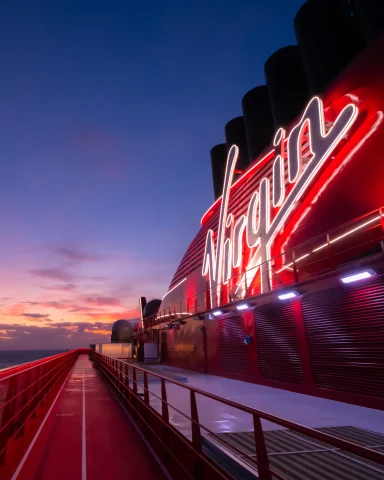 A view of a large neon red sign that says "Virgin" over the deck of a ship. There is a pink, blue and orange sunset in the distance. 