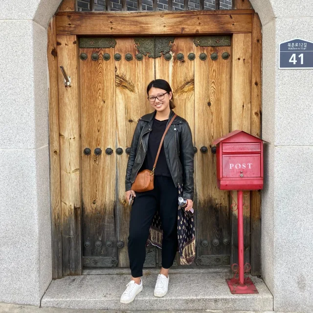 Travel Advisor Tina Chen in all black in front of a wooden door and red mailbox.