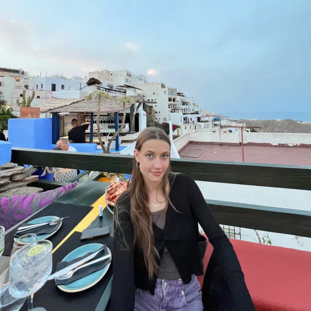 Kamila Gomez in a black cardigan sitting at a dining table on a balcony overlooking a city with white buildings