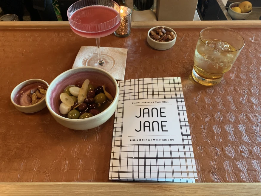 A menu at a bar reads, "Jane Jane" with a pink cocktail, a whiskey on ice, and olives and other snacks beside it