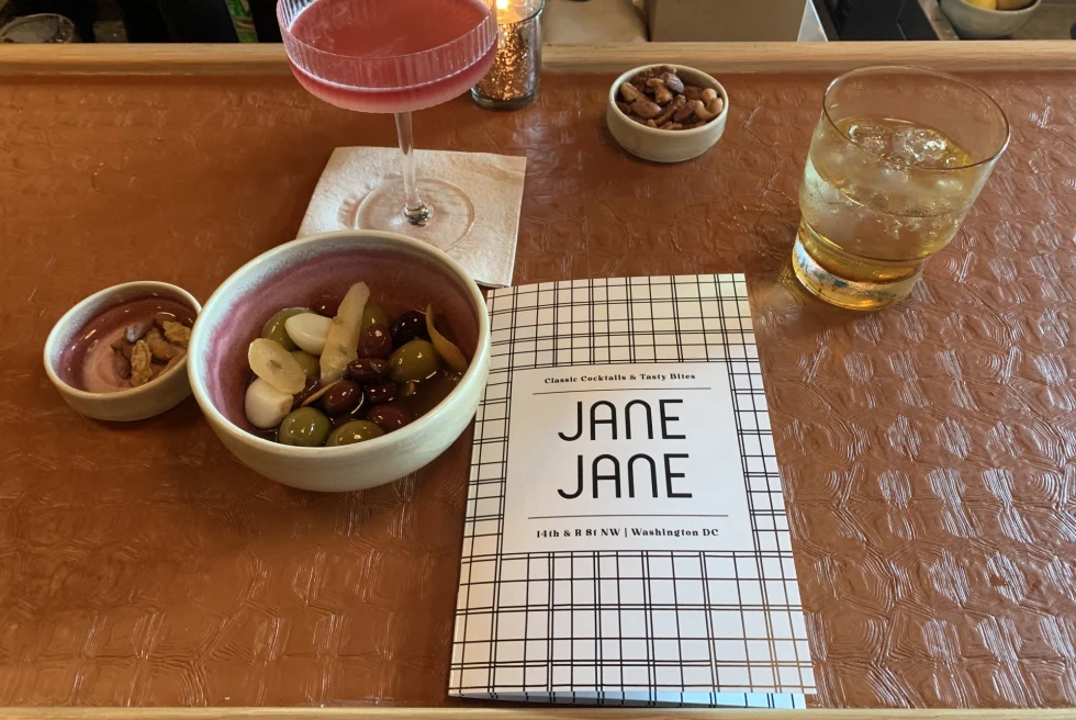 A menu at a bar reads, "Jane Jane" with a pink cocktail, a whiskey on ice, and olives and other snacks beside it