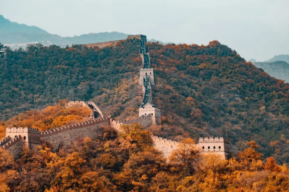 Trees with orange leaves surround the Great Wall of China on the top of a mountain with grey skies