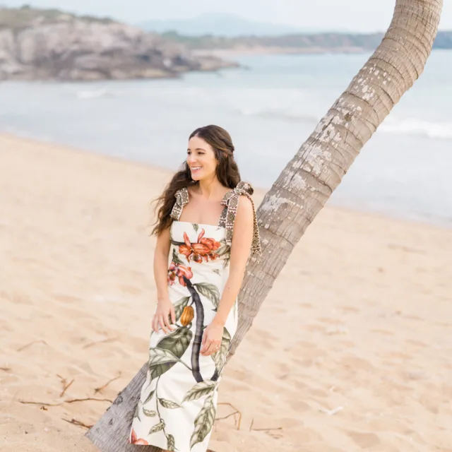 woman in a long white dress leaning against a palm tree on a beach