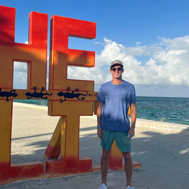 Picture of Tommy on a beach next to a red and yellow sign that says 'HE'