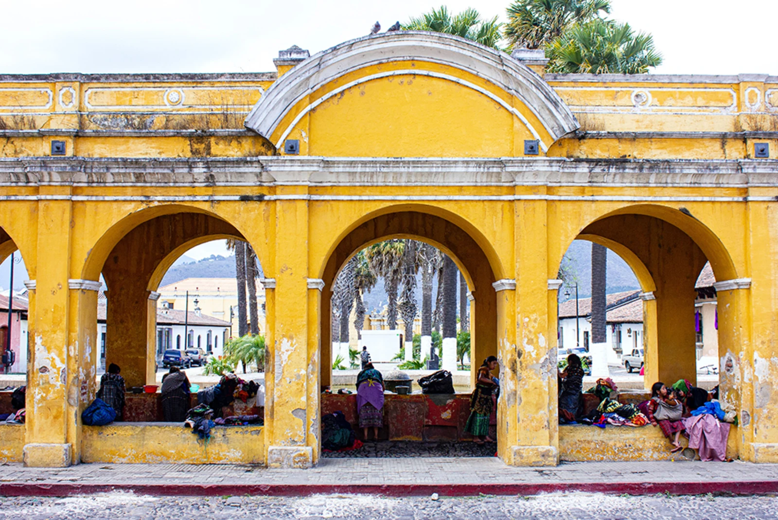 yellow and white building in Guatemala with people sitting on arches and green palm trees in background
