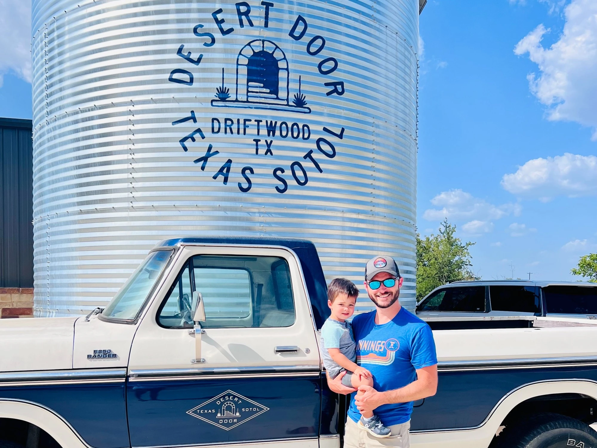 A man and kid in front of  a white and blue truck and a place saying Desert Door.