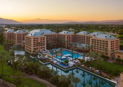 An aerial view of a resort surrounded by palm trees, a pool with lounge chairs and umbrellas, a bar a body of water and mountains beneath a golden sunset in the distance. 