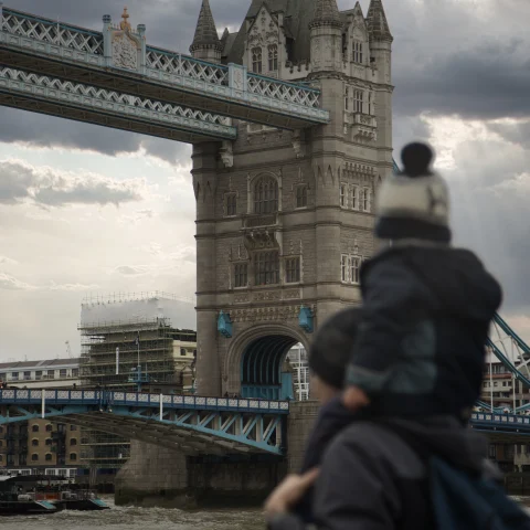 Man with child on shoulders walks past bridge and building on cloudy day