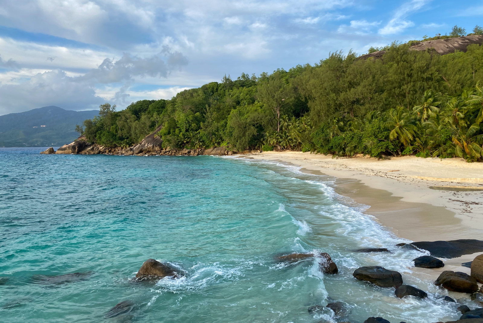 Crystal blue water with surfing waves crashing on tan sand and green lush forest in the Seychelles.
