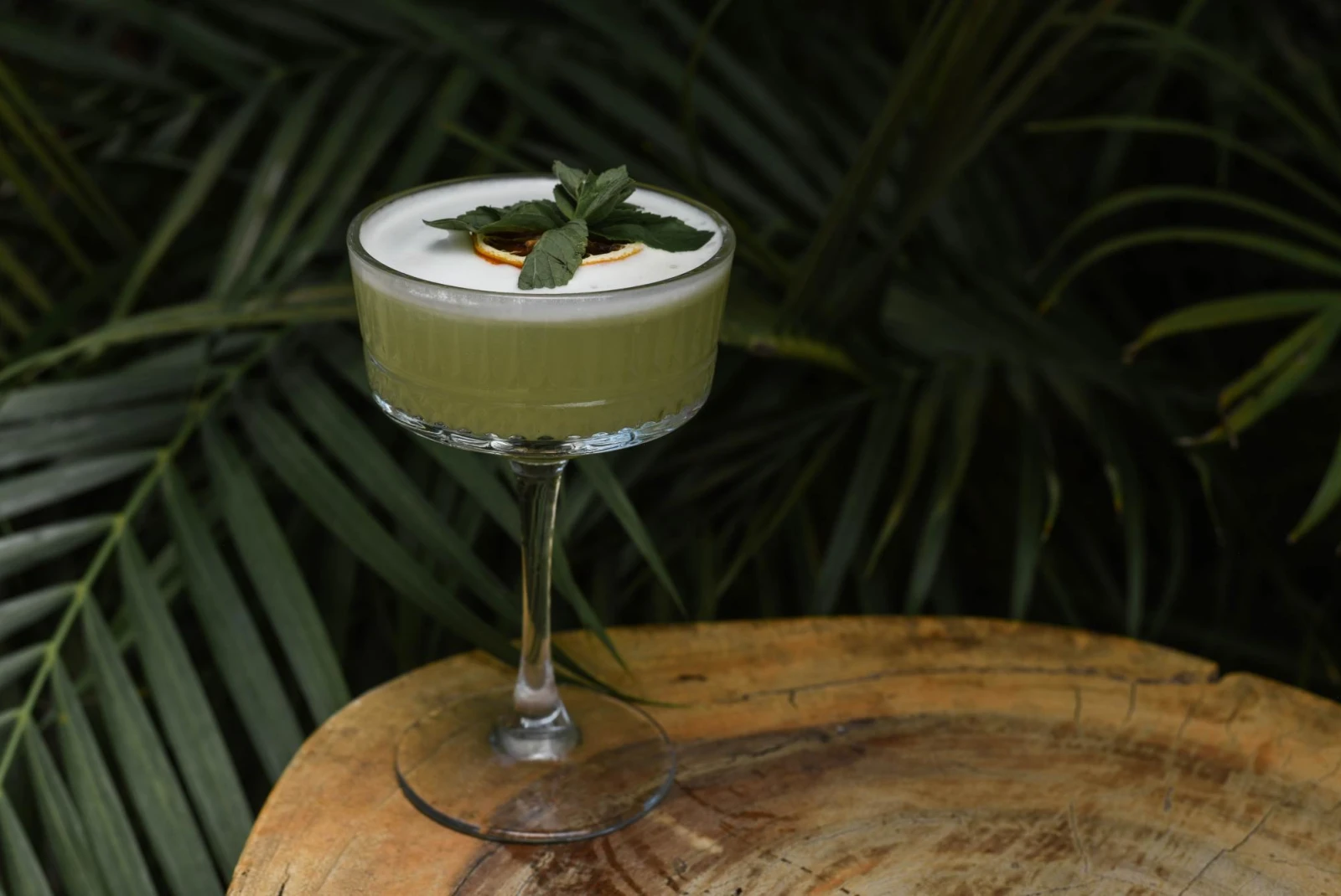 green frothy cocktail in a stemmed glass sitting on a wood table