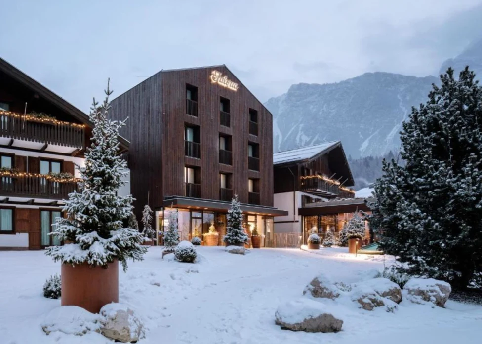 The exterior of the wooden Faloria Mountain Spa Resort on a snowy day, with green trees and mountains surrounding it.