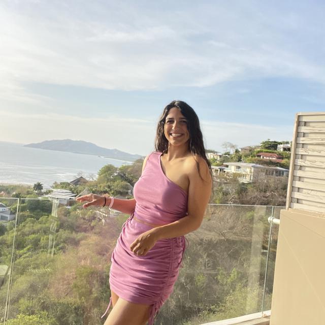 Fora travel agent Tali Gonzalez wearing pink dress standing on balcony overlooking green hills and ocean