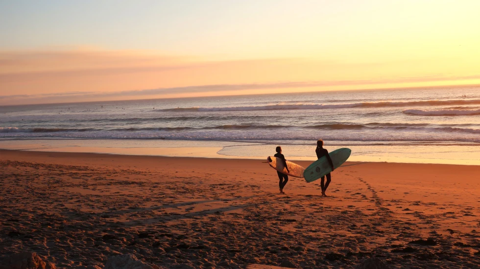 Two surfers carrying surfboards at sunset in Portugal with golden sand and pink yellow clouds.