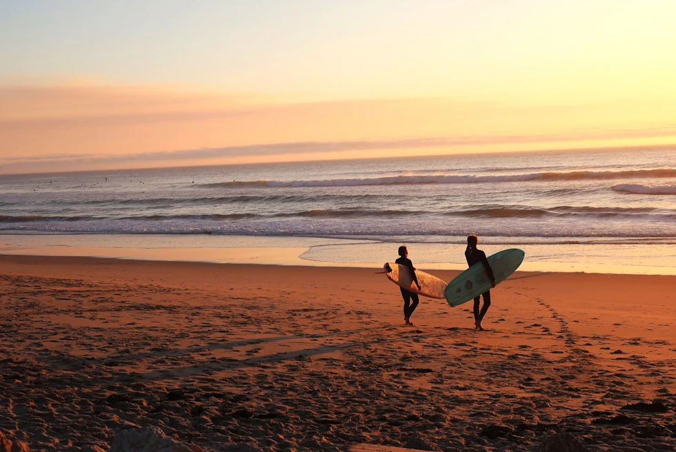 Two surfers carrying surfboards at sunset in Portugal with golden sand and pink yellow clouds.