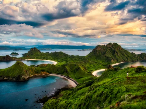 Aerial view of three separate small bays in Komodo National Park with sand in each and green hills surrounding them and clouds ahead