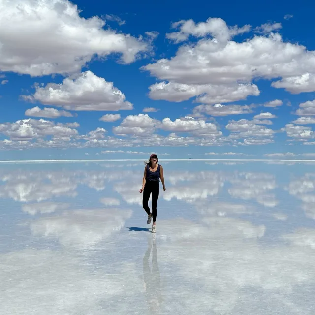 Travel Advisor Logan Hitchcock stands in shallow water with blue sky and clouds reflecting perfectly into the water