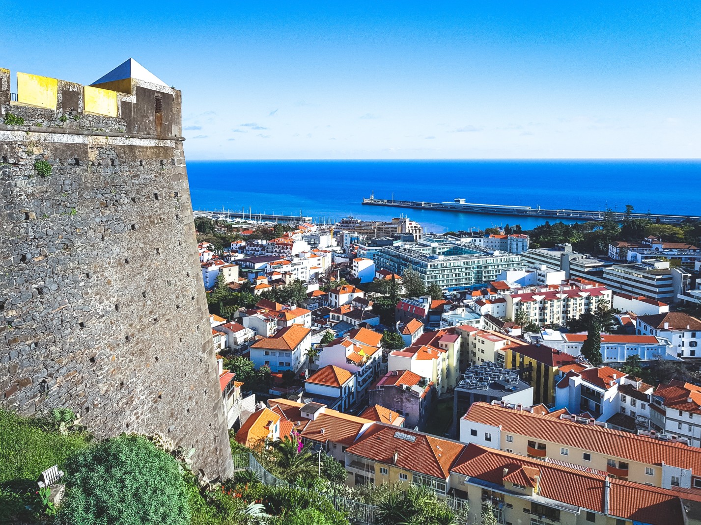 A grey stone castle-like structure overlooking a city of buildings with orange roofed buildings and a blue ocean in Funchal the capital of Madeira, Portugal. 