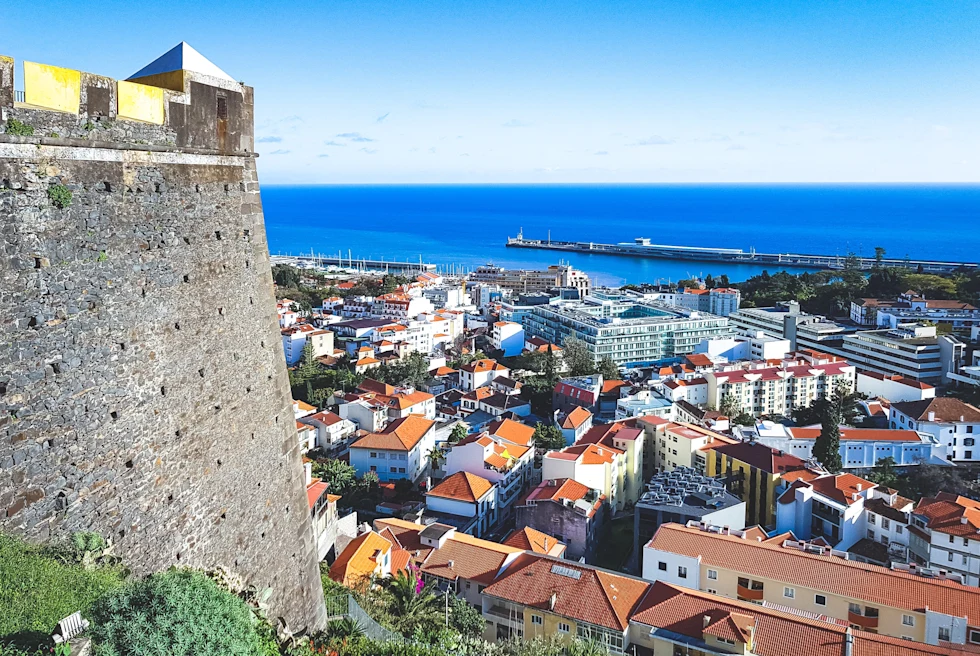A grey stone castle-like structure overlooking a city of buildings with orange roofed buildings and a blue ocean in Funchal the capital of Madeira, Portugal.